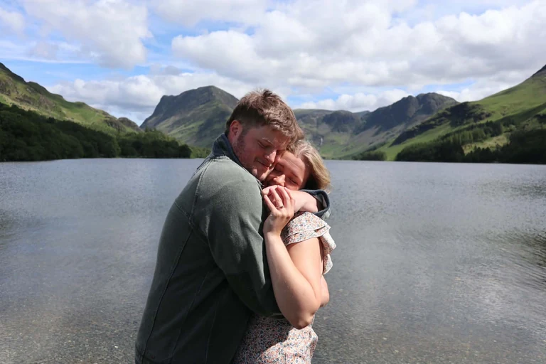Brad and Naomi holding each other by a lake, with mountains behind them