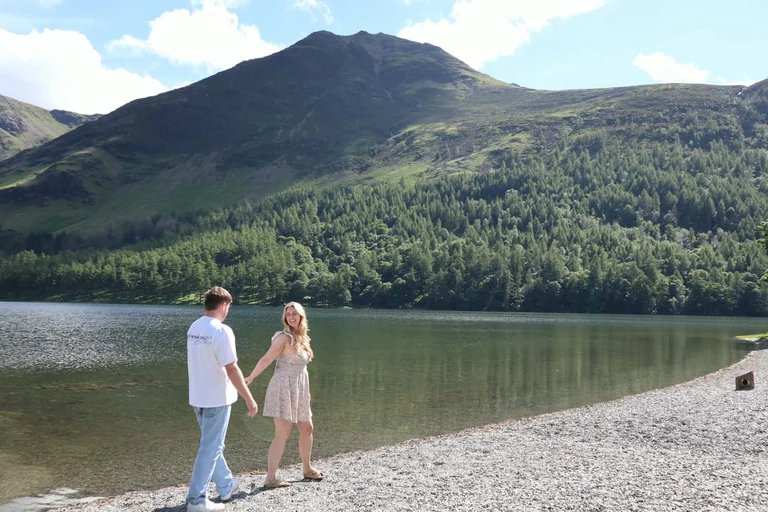 Brad and Naomi walking hand in hand along a lake shore, Naomi looking back and laughing, with a green fell rising behind them